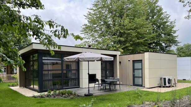 En moderne bygning med store glasfacader, en patio med udendørs siddepladser, og et lille haveområde.