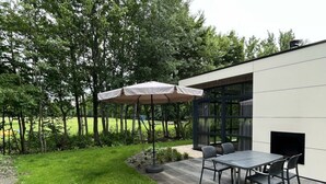 En udendørs terrasse med et bord og stole, en overdækket parasol samt en moderne bygning med store vinduer.