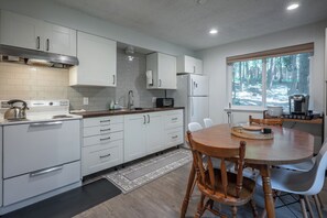 A modern kitchen with white cabinets, a stove, a microwave, and a refrigerator. There is a dining table with chairs and a window with a view of trees. - Bigfoot Escape | Dorrington Cabin with Bonus Room (Camp Connell)