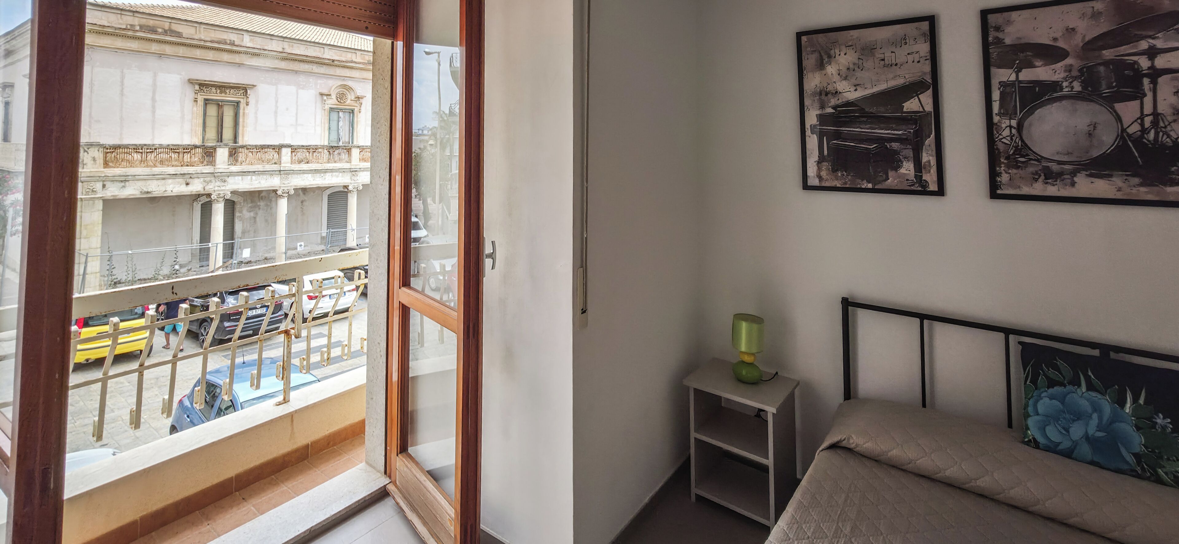 Une chambre avec un balcon, un lit, une table de chevet et des tableaux encadrés au mur.