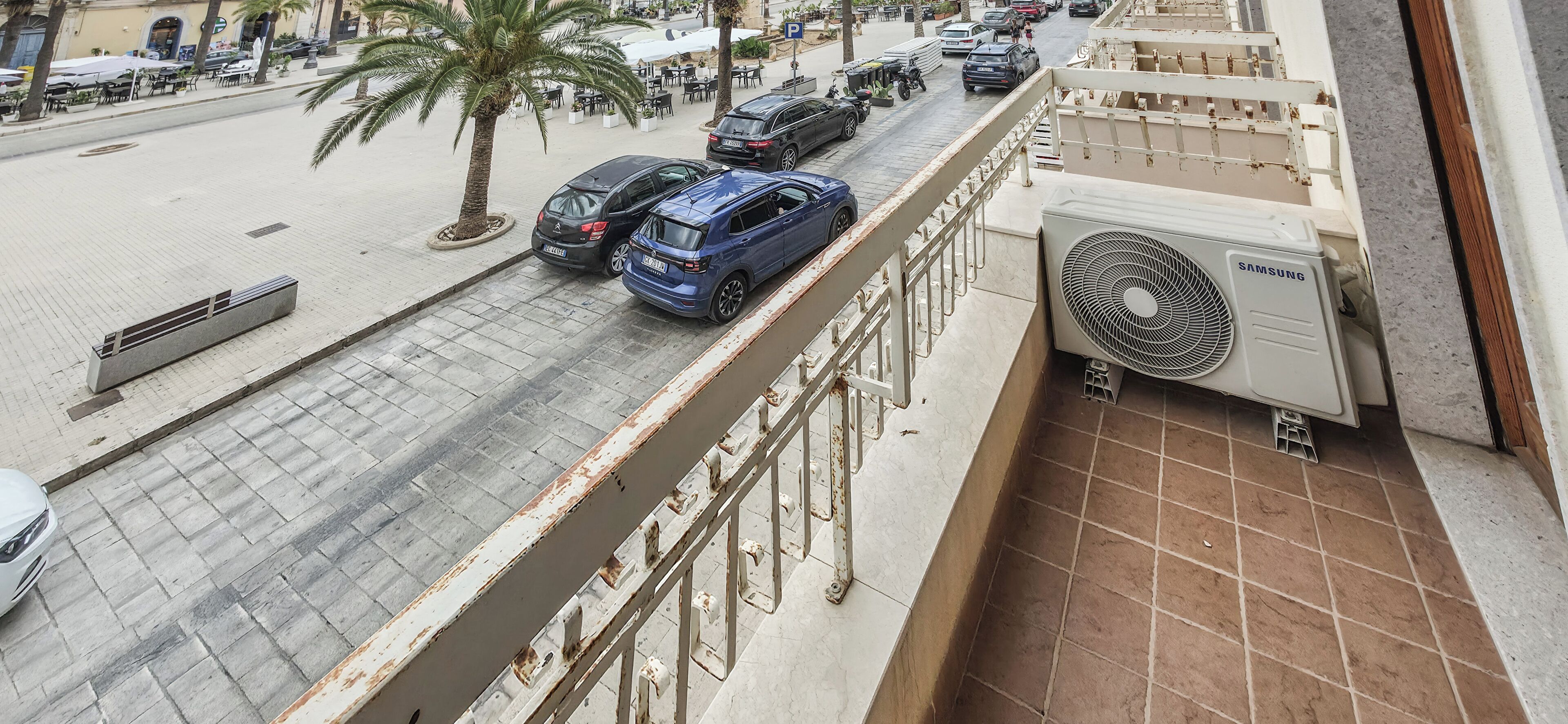 A street view with parked cars, a pedestrian walkway, and a building with a Samsung air conditioning unit.