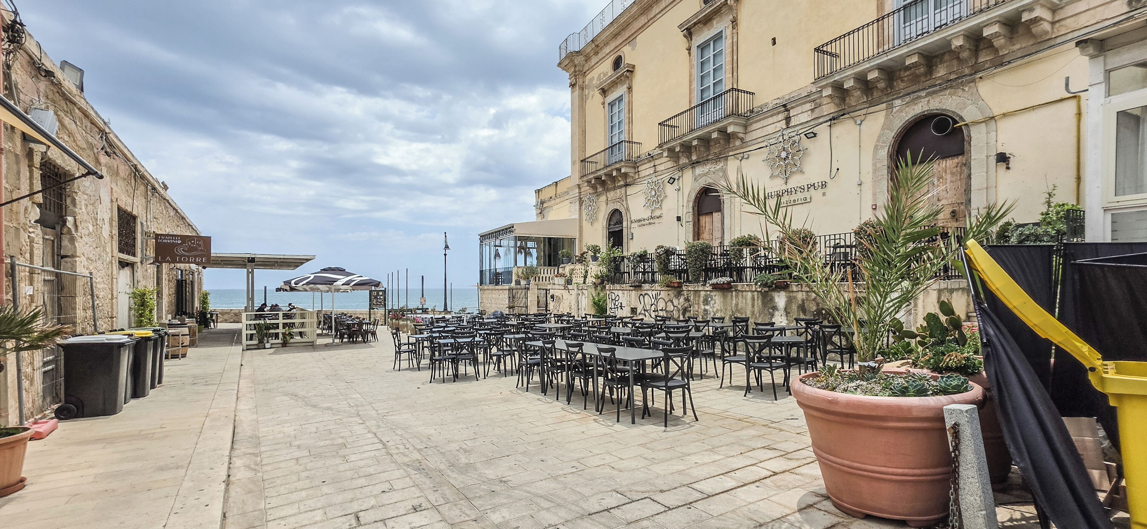 Area caffè all'aperto con tavoli e sedie, vista sul mare e un edificio con insegna.
