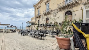 Un área de café al aire libre con mesas y sillas, vista al mar y un edificio con un cartel.