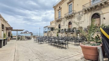 Area caffè all'aperto con tavoli e sedie, vista sul mare e un edificio con insegna.
