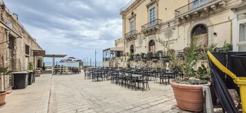 Un área de café al aire libre con mesas y sillas, vista al mar y un edificio con un cartel.