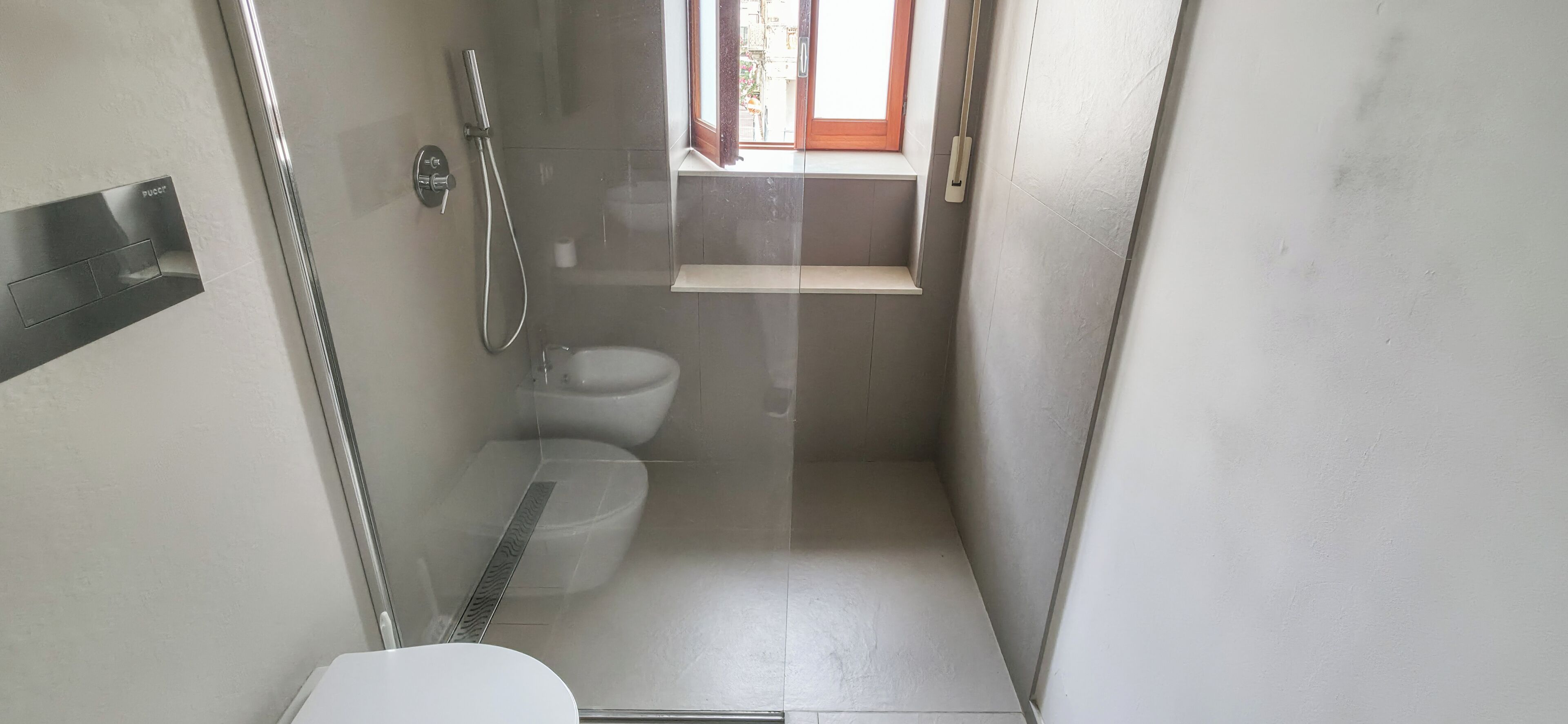 A small, narrow bathroom with a toilet, a shower, and a window.