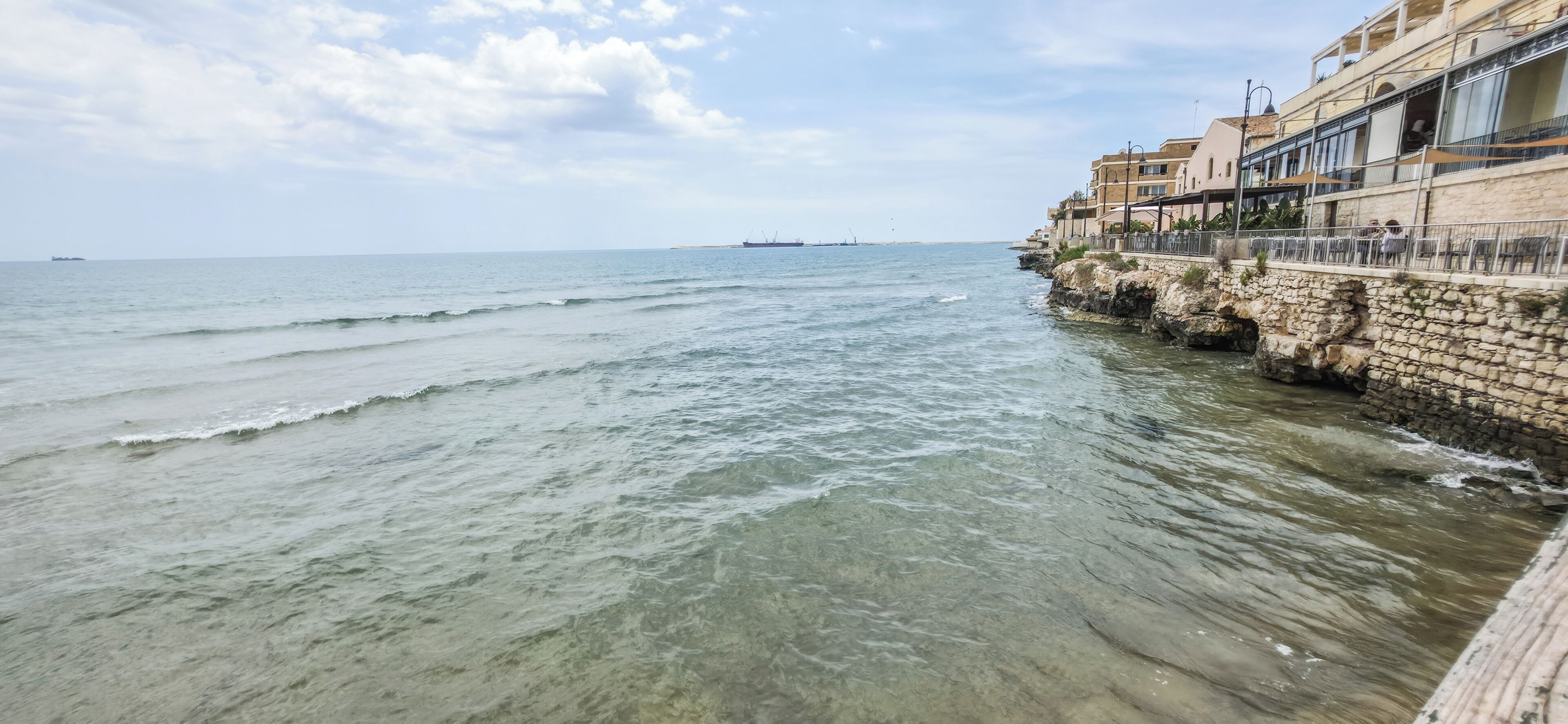 A coastal view with a stone sea wall, a building with balconies, and a distant ship.