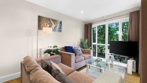 A modern living room with a sofa set, a glass coffee table, a flat-screen TV, and a large window with curtains. - Heathrow Living 2BDR 2BA Apt 03-360stays (Stanwell)