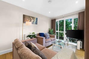 A modern living room with a sofa set, a glass coffee table, a flat-screen TV, and a large window with curtains. - Heathrow Living 2BDR 2BA Apt 03-360stays (Stanwell)