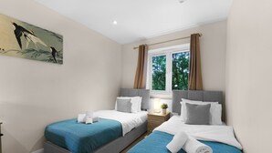 A hotel room with two beds, a window with curtains, a painting on the wall, and a small table with a lamp. - Heathrow Living 2BDR 2BA Apt 03-360stays (Stanwell)