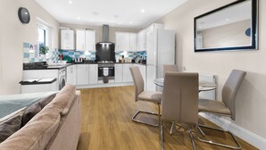 A modern kitchen with white cabinets, a black range hood, and a dining area with a glass table and chairs. - Heathrow Living 2BDR 2BA Apt 03-360stays (Stanwell)