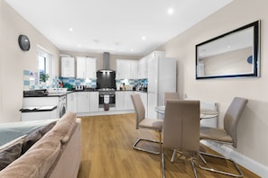 A modern kitchen with white cabinets, a black range hood, and a dining area with a glass table and chairs. - Heathrow Living 2BDR 2BA Apt 03-360stays (Stanwell)