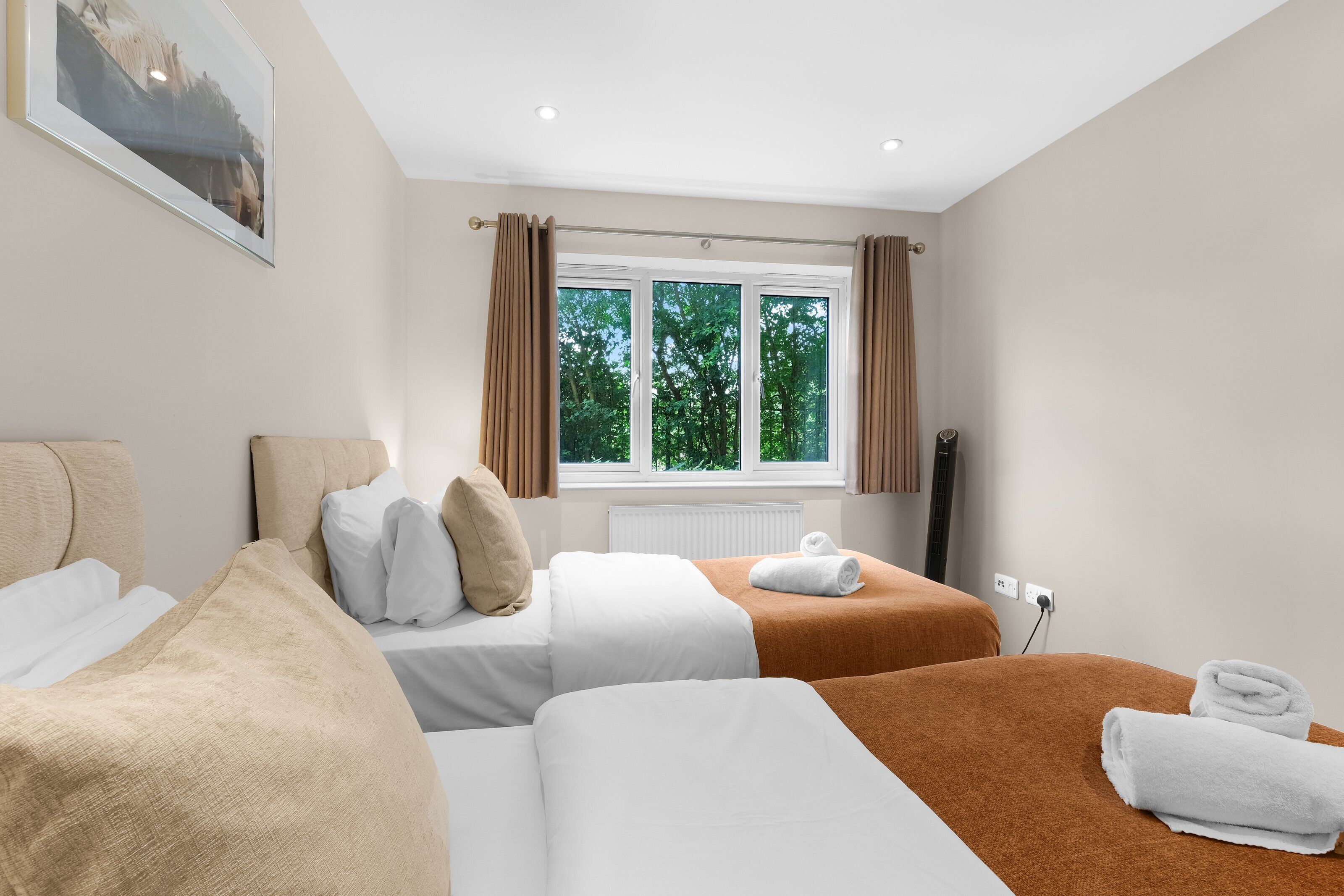 A hotel room with two beds, a window with curtains, a radiator, and a power outlet.