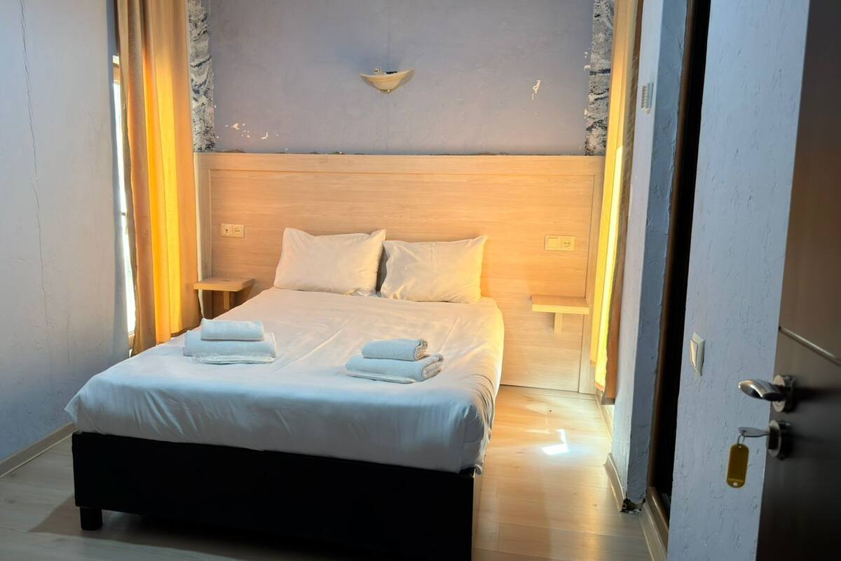A neatly made bed with white linens and towels, a wooden headboard, and a wall-mounted light fixture.