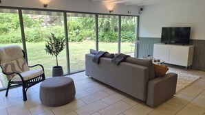 Un salon moderne comprenant un canapé, un pouf et un fauteuil, un téléviseur et une grande porte-fenêtre coulissante donnant sur un jardin.