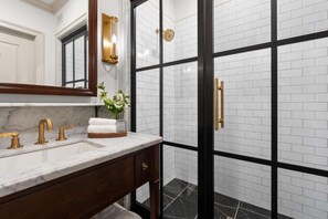 A bathroom with a marble countertop, a gold faucet, a glass vase with flowers, and a black and white tiled shower area.