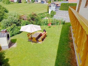 Ein Garten mit Terrasse, Tisch, Sonnenschirm, Schaukel und einem kleinen Spielbereich.