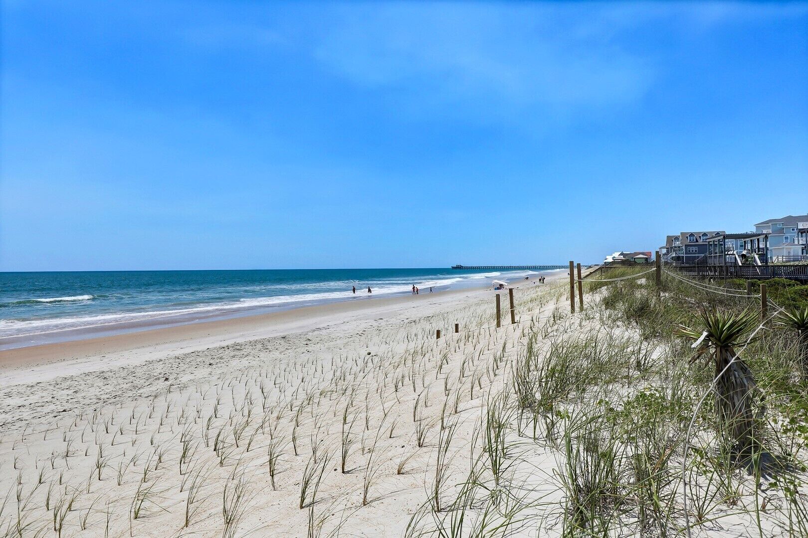 A beach with a wooden fence, a pier in the distance, and buildings on the right.