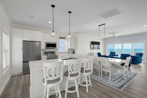 A modern kitchen with white cabinetry, a central island with bar stools, a dining area with blue chairs, and a large window overlooking the ocean. - Oceanfront Luxury Retreat | Private Beach Access | Sunrise & Sunset Views (North Topsail Beach)