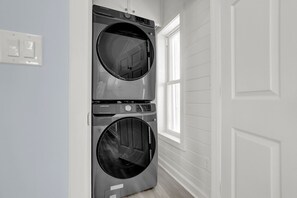 A laundry room with two stacked front-loading washers and dryers, a window, and a white door. - Oceanfront Luxury Retreat | Private Beach Access | Sunrise & Sunset Views (North Topsail Beach)