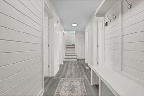 A hallway with white paneling, a patterned rug, and a staircase leading to another room. - Oceanfront Luxury Retreat | Private Beach Access | Sunrise & Sunset Views (North Topsail Beach)
