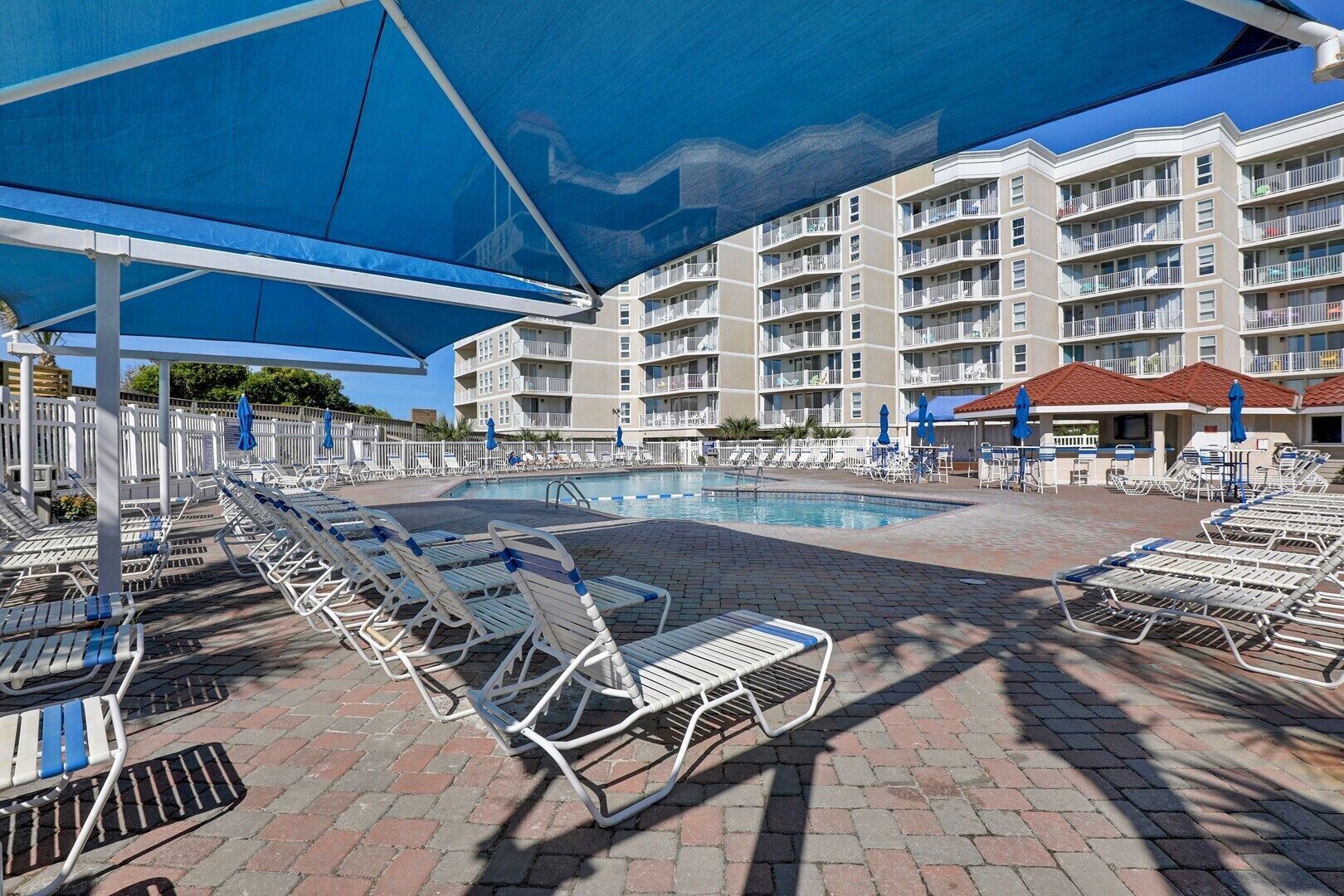 A pool area with lounge chairs and umbrellas, a pool, and a multi-story building with balconies.
