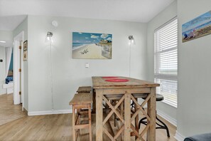 Ein Zimmer mit einem Holztisch, zwei Wandleuchten, einem Strandbild und einem Fenster mit Jalousien.