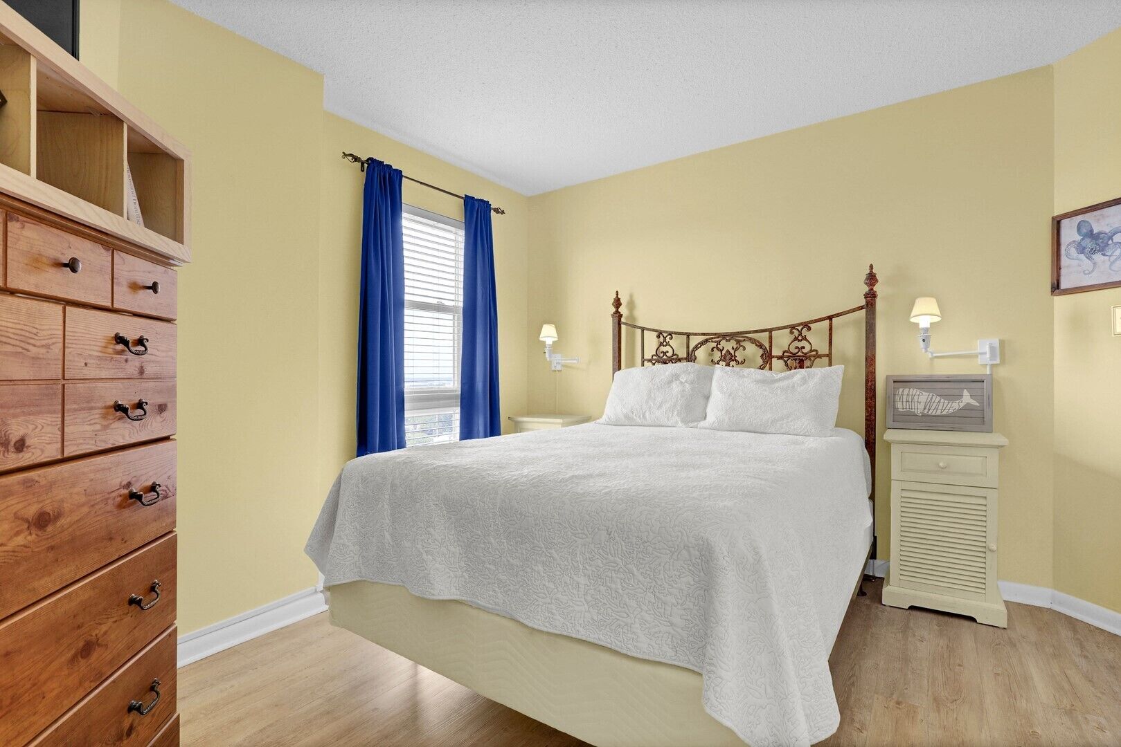 Un dormitorio con cómoda de madera, cama con ropa blanca, mesita de noche con lámpara y un cuadro colgado en la pared.