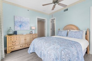 A bedroom with a bed, dresser, ceiling fan, and a door leading to another room.