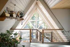 A room with a sloped ceiling, a wooden staircase, and a large window with a view of trees. - Marmot Cabin | Cedar Tub, Fireplace, Pets (Packwood)