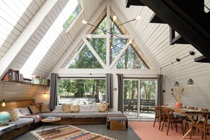 A cozy loft-style room with a sloped ceiling, built-in seating, a dining area, and large windows offering a view of the outdoors. - Marmot Cabin | Cedar Tub, Fireplace, Pets (Packwood)