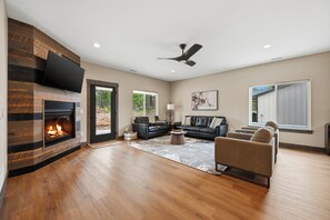 A spacious living room with a fireplace, a flat-screen TV mounted on a wooden wall, a ceiling fan, and a large window.