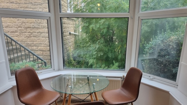 Habitación de esquina con dos sillones marrones, una mesa de vidrio y ventanas amplias que dan a zonas verdes.