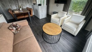 A modern living room with a sofa, two armchairs, a coffee table, a dining area with a table and chairs, a flat-screen TV, and a view of greenery outside. - Holiday Home in Susteren With Sauna (Susteren)