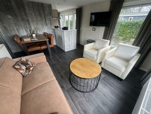 Een moderne woonkamer met een bank, twee fauteuils, een salontafel, een eethoek met tafel en stoelen, een flatscreen-tv en uitzicht op groen.