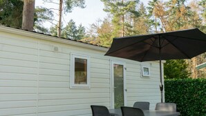 A small, single-story mobile home with a patio area featuring a table and chairs, an umbrella, and a small window. - Holiday Home in Susteren With Sauna (Susteren)