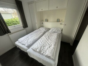 Een kleine kamer met een enkel bed, raam, radiator en een plank boven het bed.