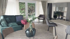 Una sala moderna con un sofá verde, una mesa redonda con un florero y una zona de cocina con televisión y armarios.