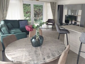 Ein modernes Wohnzimmer mit einem grünen Sofa, einem runden Tisch mit einer Vase sowie einer Küchenzeile mit Fernseher und Schränken.