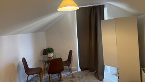 Et lille, godt oplyst rum med et glasbord, fire stole, en potteplante og et stort vindue med gardiner.