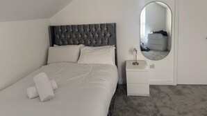 Et soveværelse med en polstret sengegavl, et natbord med en lampe, et spejl og en fint redt seng med hvide sengetøj og håndklæder.