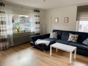 A living room with a dark blue sofa, a white coffee table, sheer curtains, a potted plant, and a radiator.