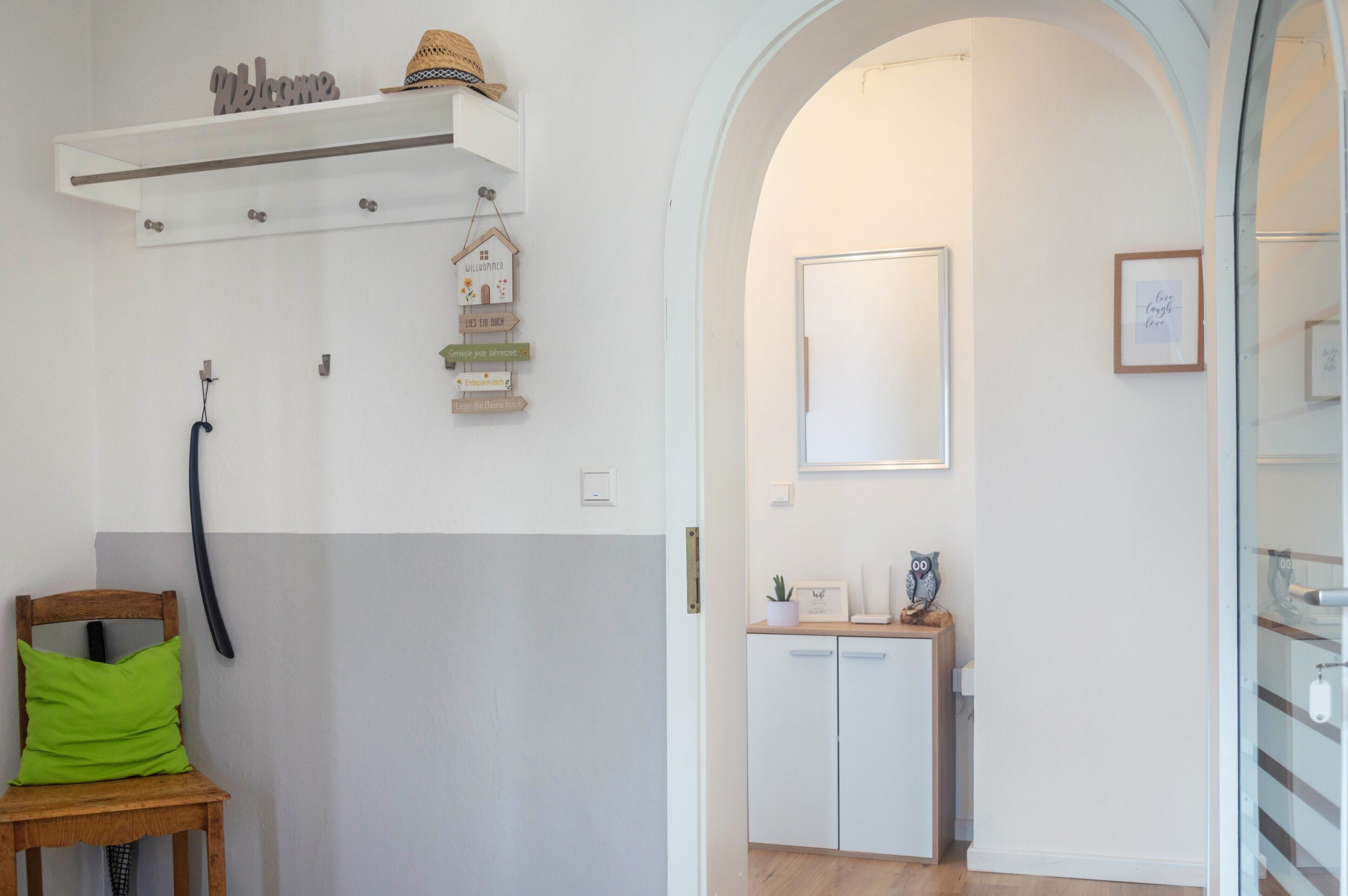 A room with a wooden chair, a white wall with a 'Welcome' sign, a small shelf with a hat, and a door leading to another room.