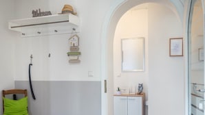 A room with a wooden chair, a white wall with a 'Welcome' sign, a small shelf with a hat, and a door leading to another room.