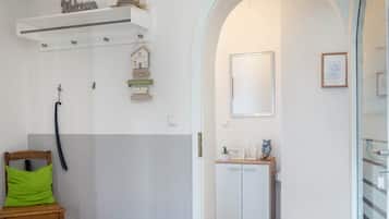 A room with a wooden chair, a white wall with a 'Welcome' sign, a small shelf with a hat, and a door leading to another room.