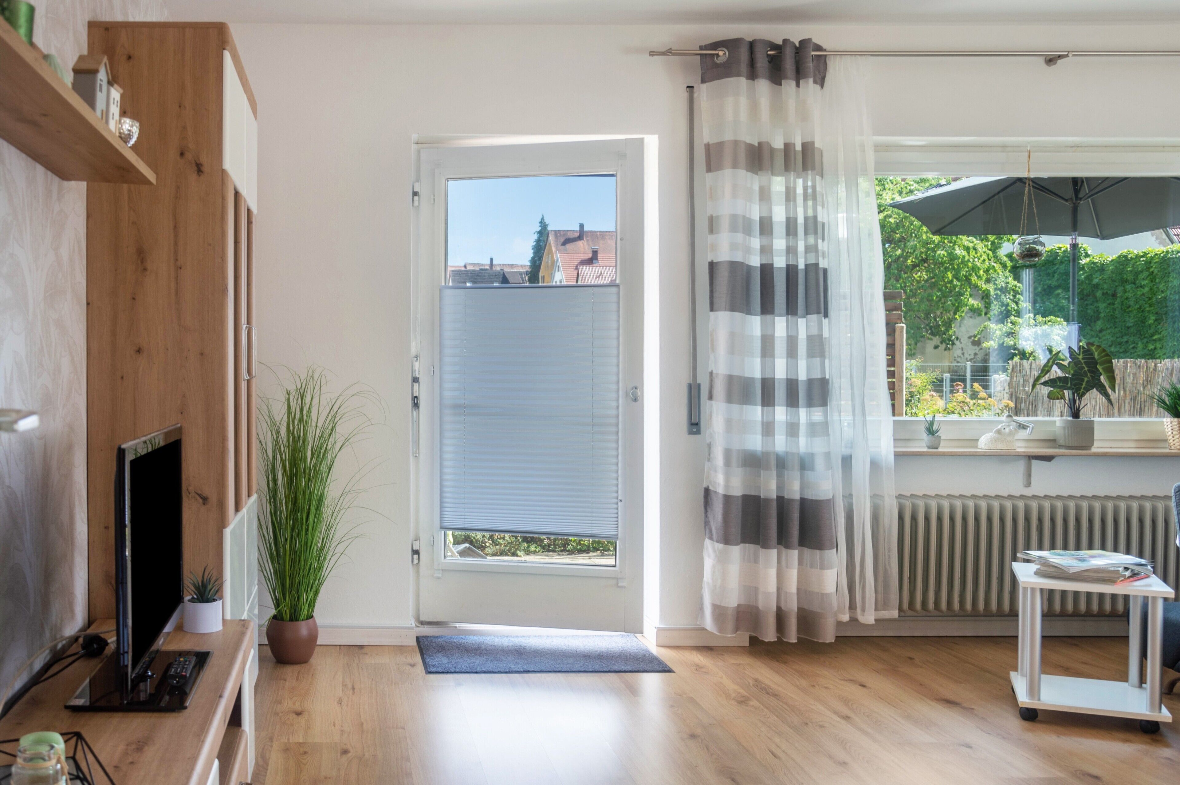 Een kamer met houten vloer, een televisie, een kamerplant, een deur met uitzicht, een radiator en een raam met gestreepte gordijnen.