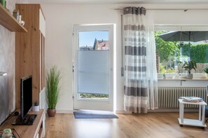 Une pièce avec un plancher en bois, un téléviseur, une plante en pot, une porte donnant sur une vue agréable, un radiateur et une fenêtre avec un rideau à rayures.