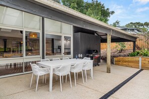 A modern outdoor dining area with a white table and chairs, a grill, and a view of the interior through large windows. - Modern Beach holiday House in Rhyll close to Beach, Restaurants, boating (Rhyll)