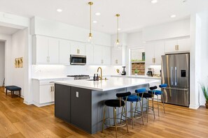 A modern kitchen with white cabinetry, a central island with bar stools, stainless steel appliances, and pendant lighting. - Golf Getaway | Pool, Spa, Arcades, BBQ, PGA West! (La Quinta)
