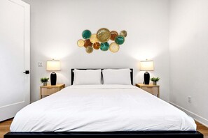 A bedroom with a large bed, two bedside tables with lamps, and a wall decoration. - Golf Getaway | Pool, Spa, Arcades, BBQ, PGA West! (La Quinta)